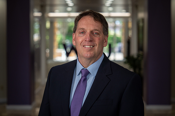 A man in a dark suit and purple tie (Chris Mallett) smiles while standing in a softly lit office hallway.