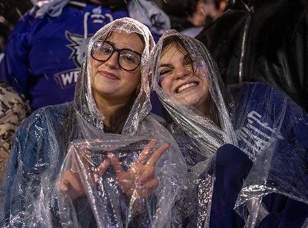 Students in the rain at a football game wearing ponchos and smiling at the camera