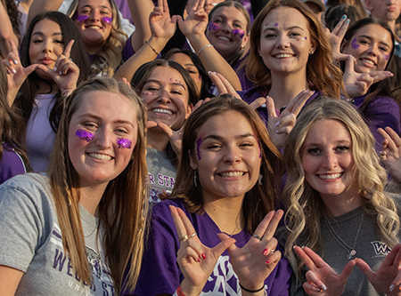Girls in the crowd at a Weber State football game, smiling at the camera making a "W" sign with their hands.