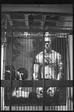 picture of tatooed man behind bars, another lifting weights in the background.